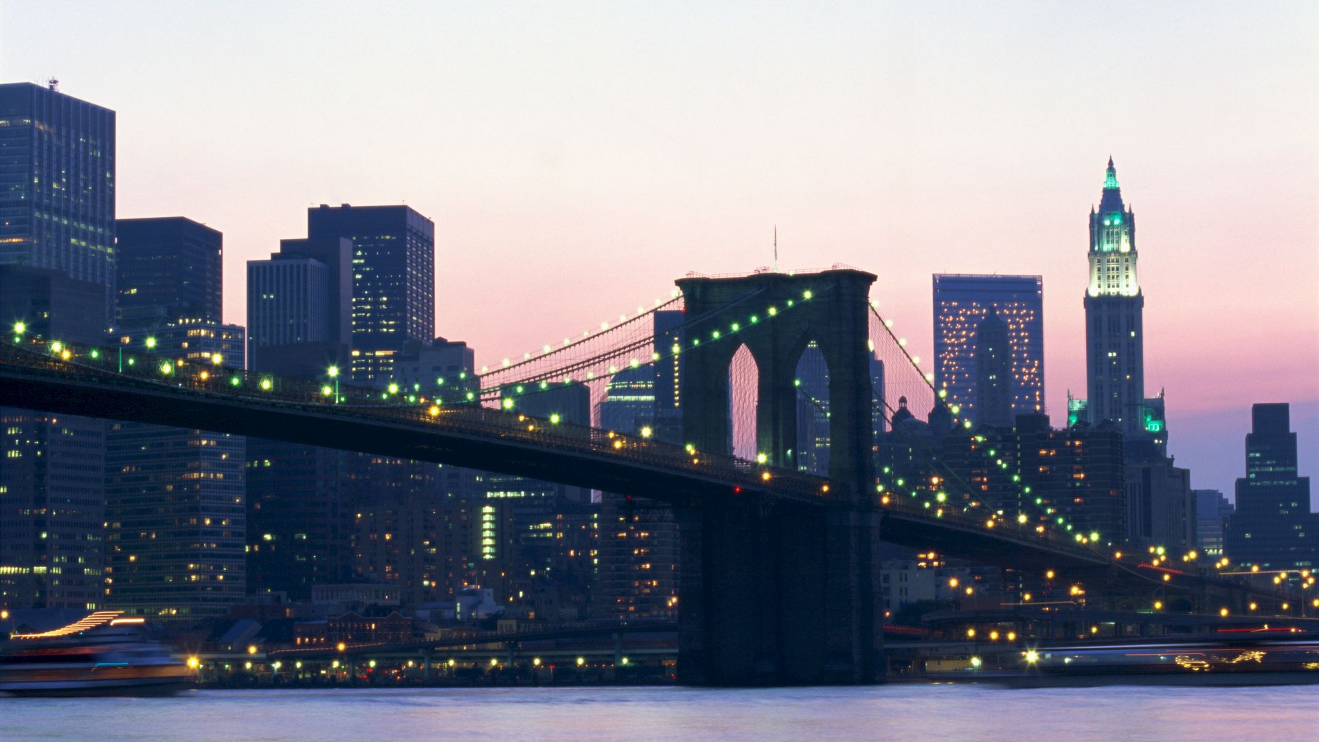A view of the Brooklyn Bridge with the New York City skyline at dusk, featuring illuminated skyscrapers and a tranquil river.
