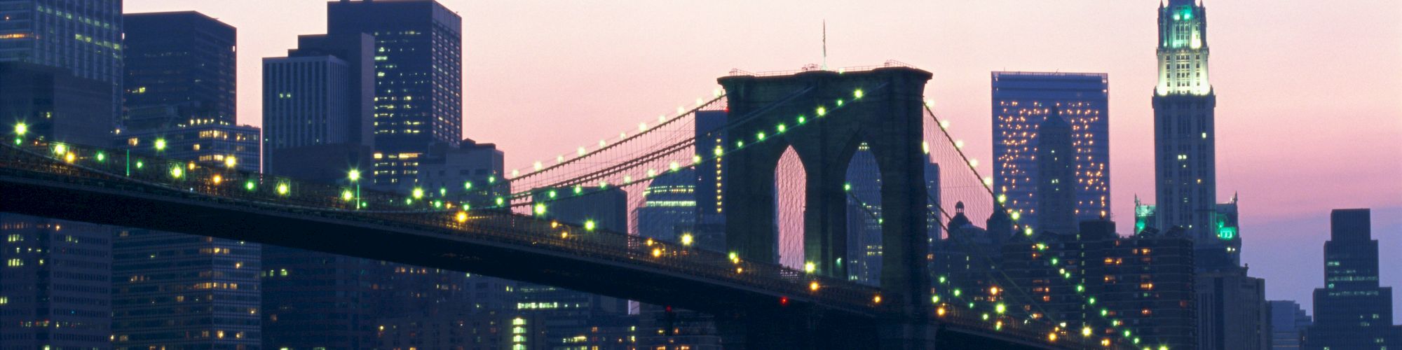 A view of the Brooklyn Bridge with the New York City skyline at dusk, featuring illuminated skyscrapers and a tranquil river.