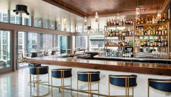 The image shows a modern bar with a wooden counter, blue bar stools, and shelves filled with various bottles. Large windows provide city views.