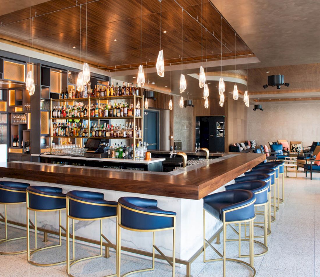 A wide view of a bar with a long counter, blue stools, and shelves of bottles.