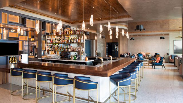 A wide view of a bar with a long wooden counter, blue stools, and pendant lights.