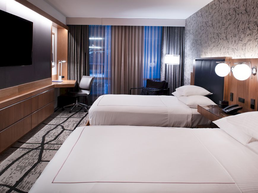 A modern hotel room with two neatly made beds, a flat-screen TV, a desk with a chair, and a stylish wall design behind the beds.