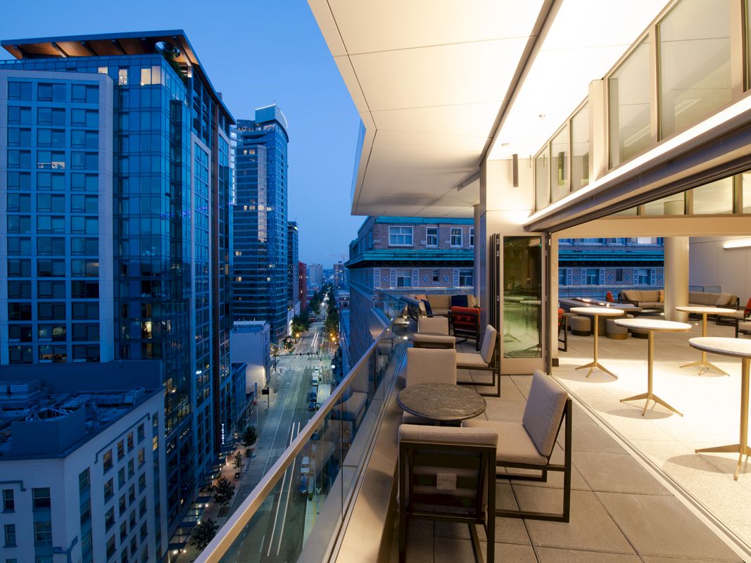 A modern, cityscape view from a balcony with seating arrangements and tables, overlooking a street flanked by tall buildings at dusk.