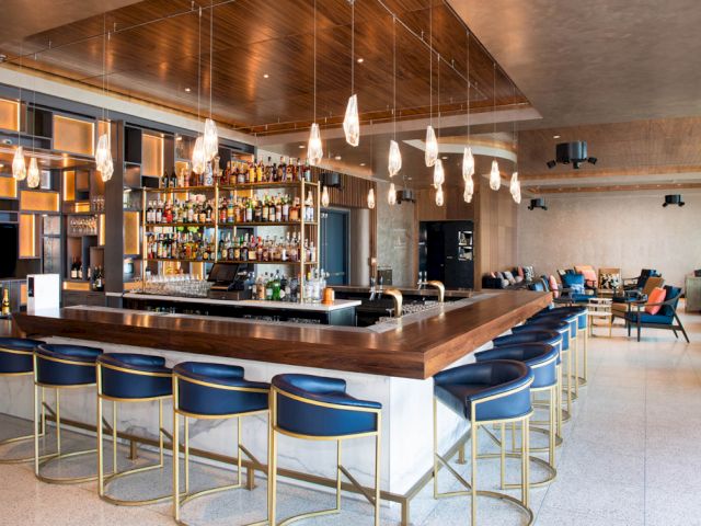 A wide view of a bar with blue stools, a wooden counter, and shelves of bottles in the background.