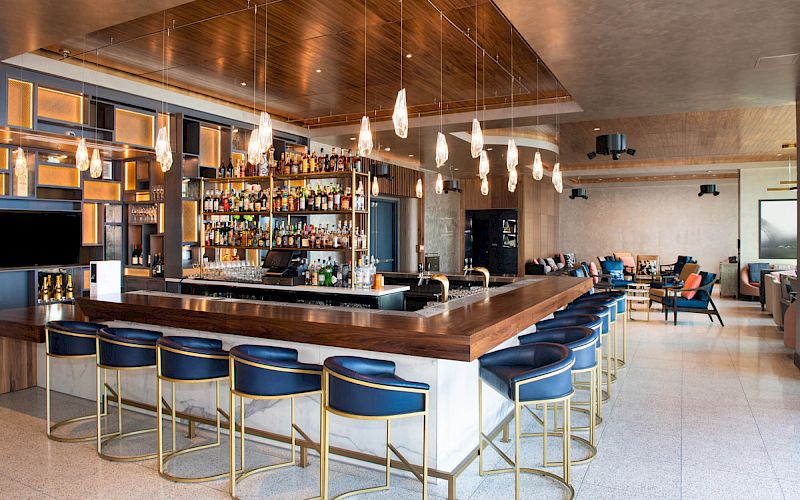 The image shows a modern bar with a wooden counter, blue stools, hanging pendant lights, and a fully stocked back wall, creating a sleek atmosphere.