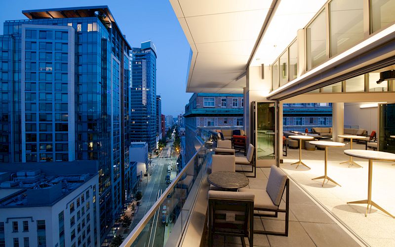 A modern cityscape viewed from a high balcony features tall buildings and a stylish outdoor seating area in the evening.