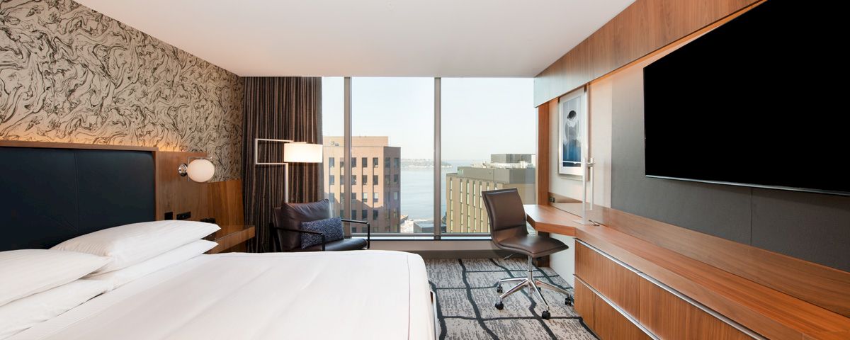 A hotel room with a large bed, desk, TV, and window with a city view.