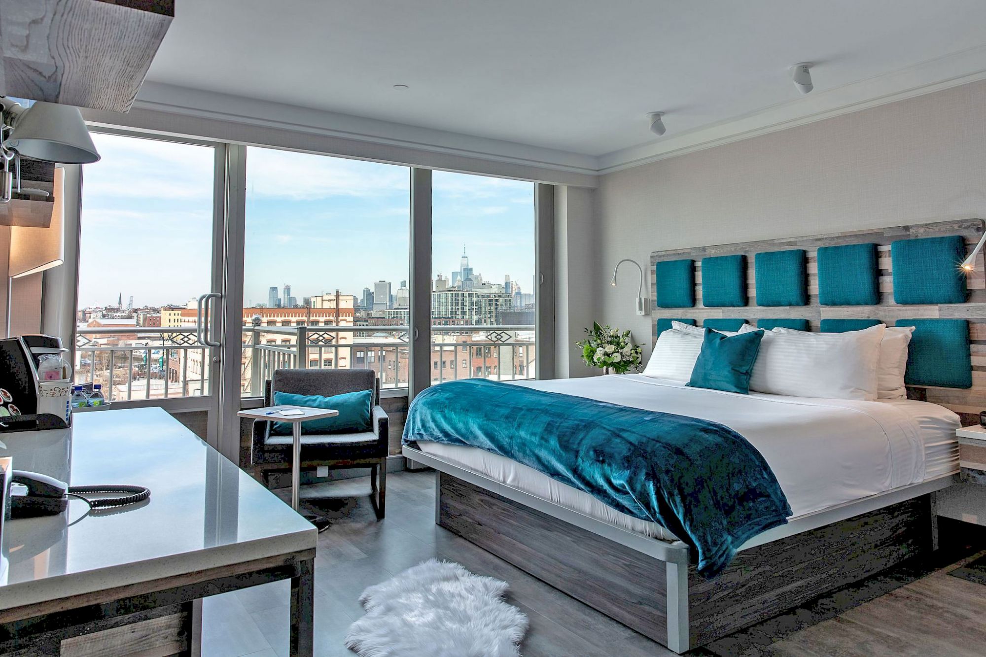 A modern hotel room with a large bed, teal accents, a desk, and a view of a city skyline through large windows, ending the sentence.