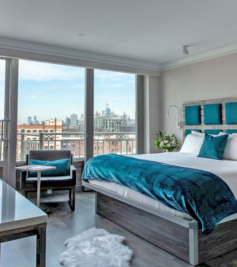 A hotel room with a bed, chair, table, and a large window showing a city view.