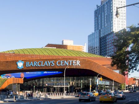 Image of Barclays Center, a modern arena with a green roof and glass front located in an urban setting with nearby buildings and street traffic.